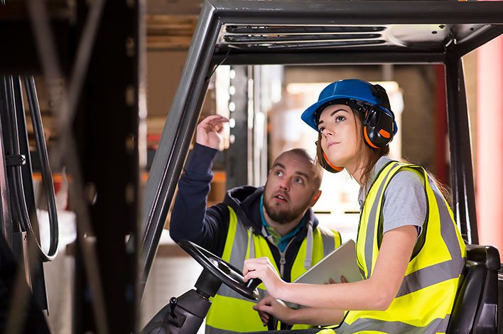 A man instructs a young woman on using a forklift.