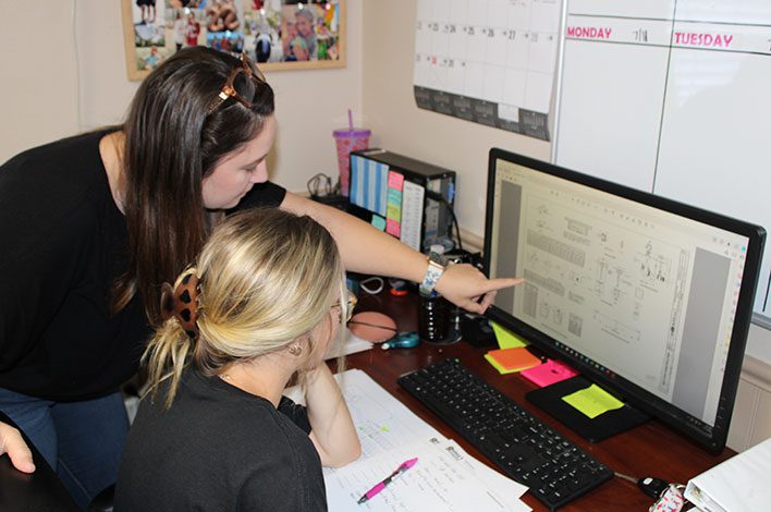 A woman leans over a young woman's shoulder to point out something on a computer screen.