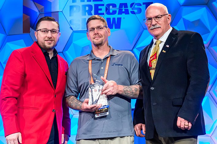 Three men stand on a stage. The man in the middle is holding a trophy.