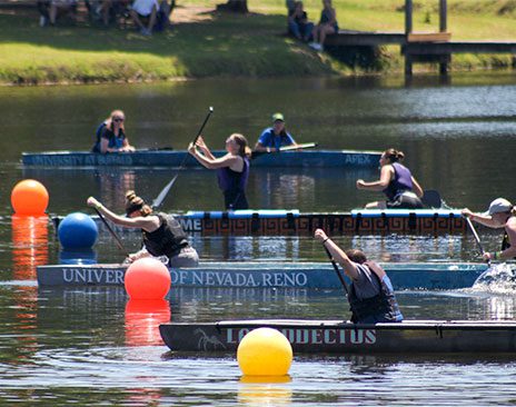 Three canoes with 2 people in each of them race to cross a finish line marked by floating balls.
