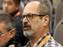 A man in glasses and a goatee speaks into a microphone.