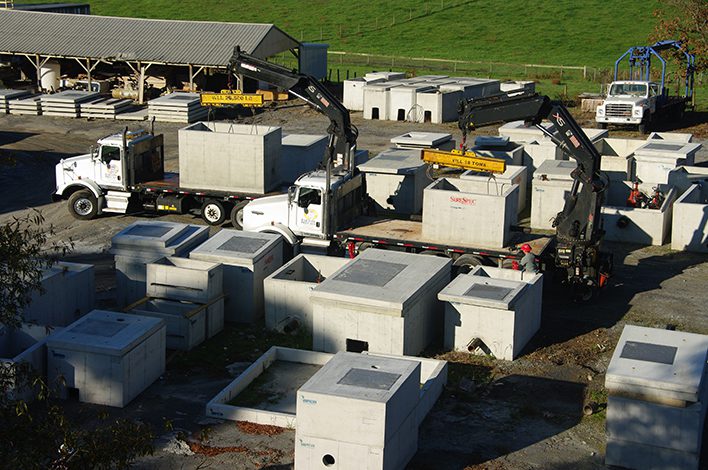Cranes load large, rectangular precast concrete products onto flatbed trucks in a precast yard.