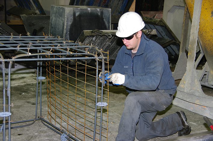 A man wearing protective equipment, including gloves, eye protection and a hardhat, works to build a rebar cage.